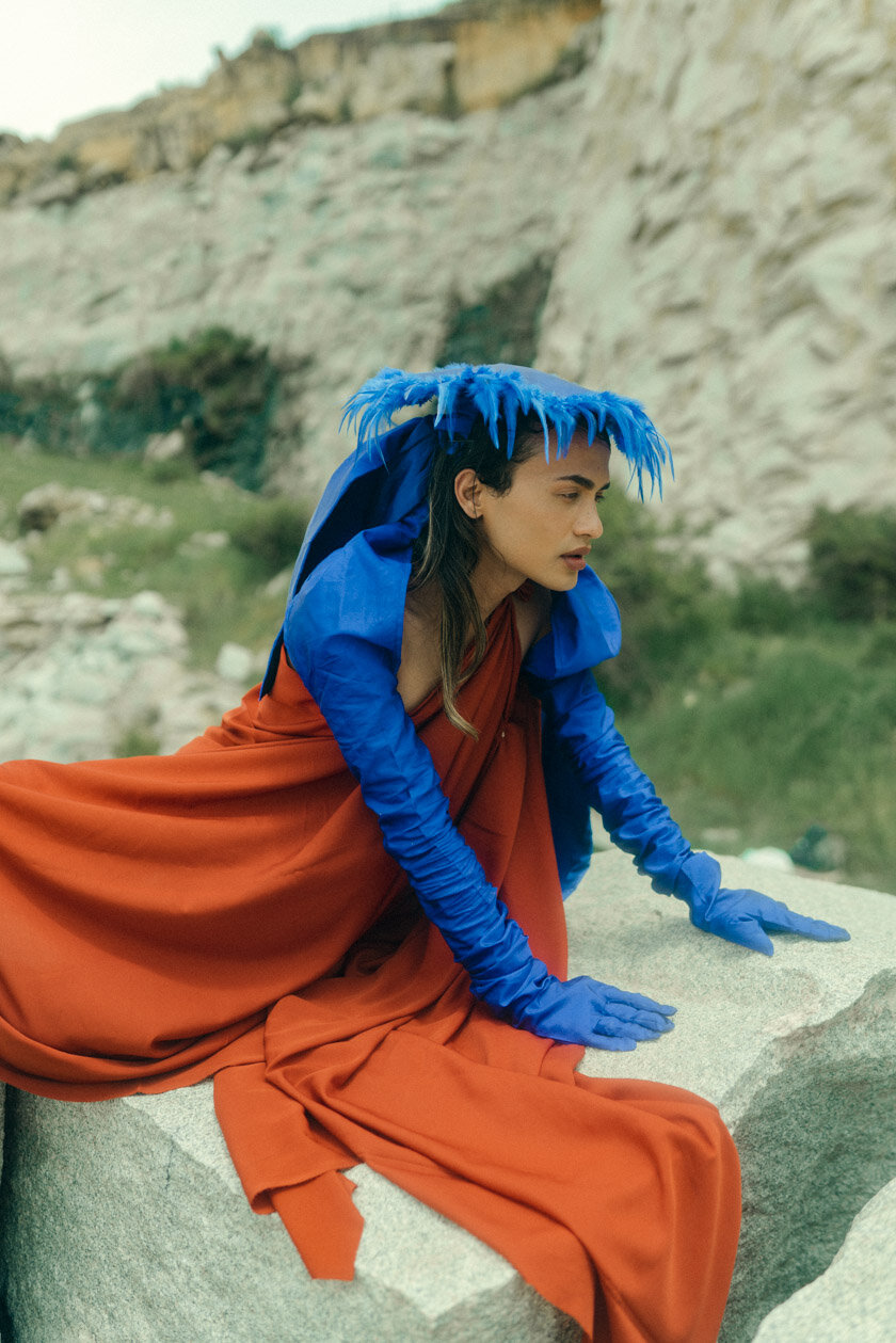 A woman dressed in a flowing orange gown, blue gloves, and a blue feathered headdress, sitting on a large rock outdoors with a rocky hillside in the background.