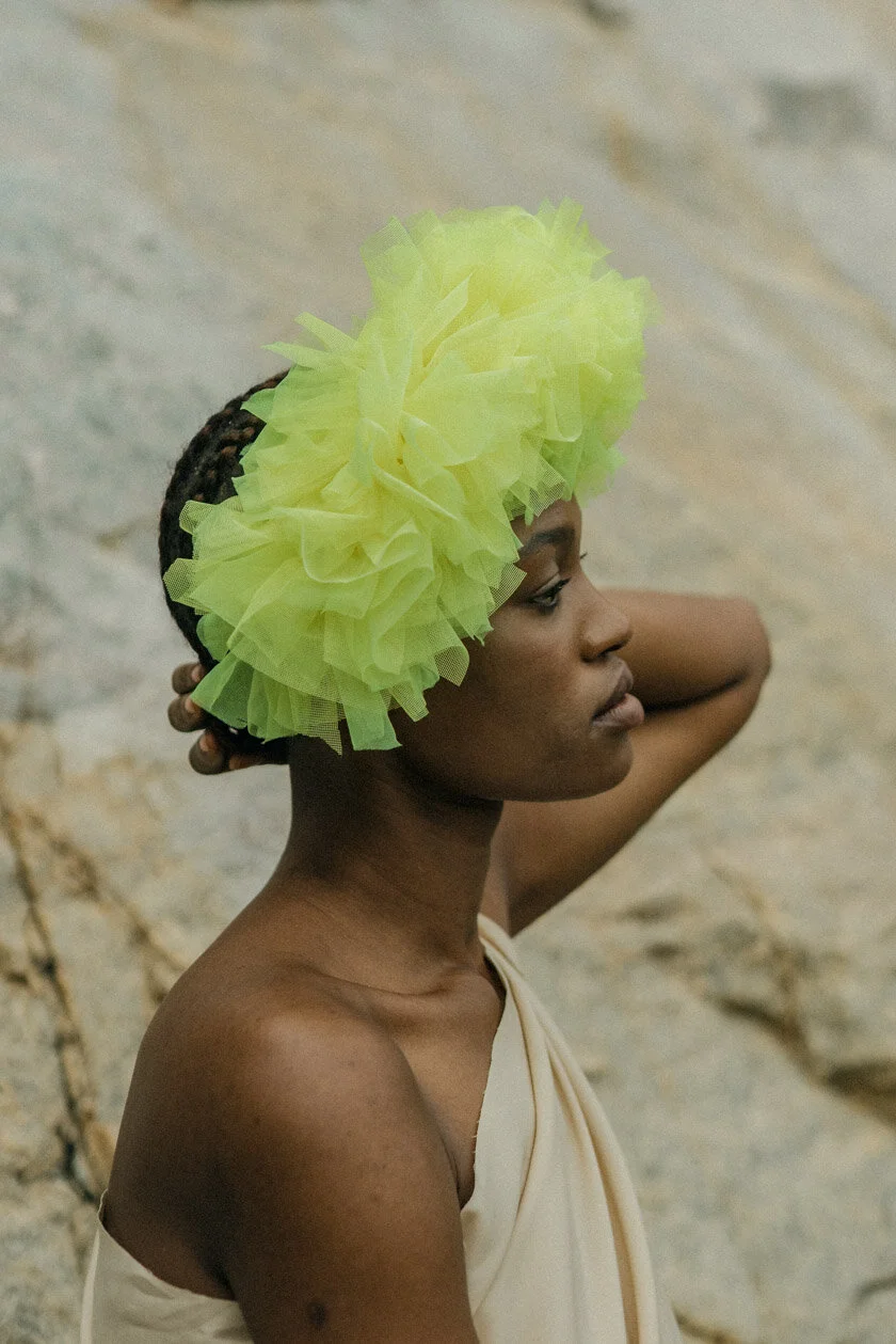 A woman with dark skin, wearing a large, light yellow tulle headpiece, poses against a rocky background, with her right arm raised and hand resting on the back of her head.