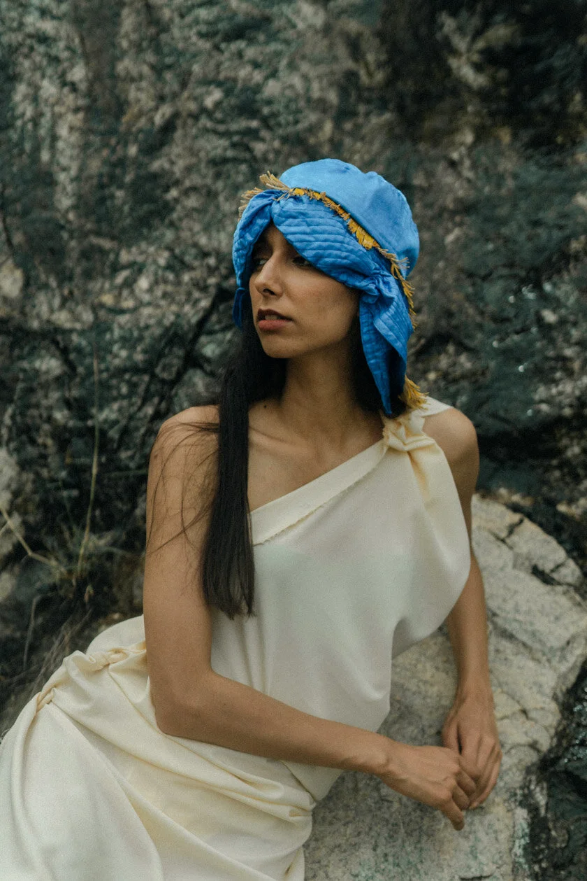 A woman with long dark hair sitting outdoors among rocks, wearing a white off-shoulder dress and a blue quilted head covering.