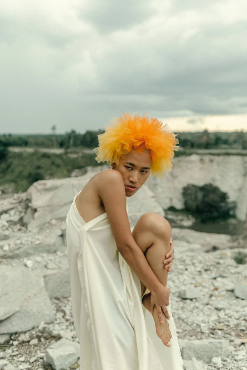 Person with a yellow feathered headpiece wearing a white dress, standing on rocky terrain with a cloudy sky in the background.