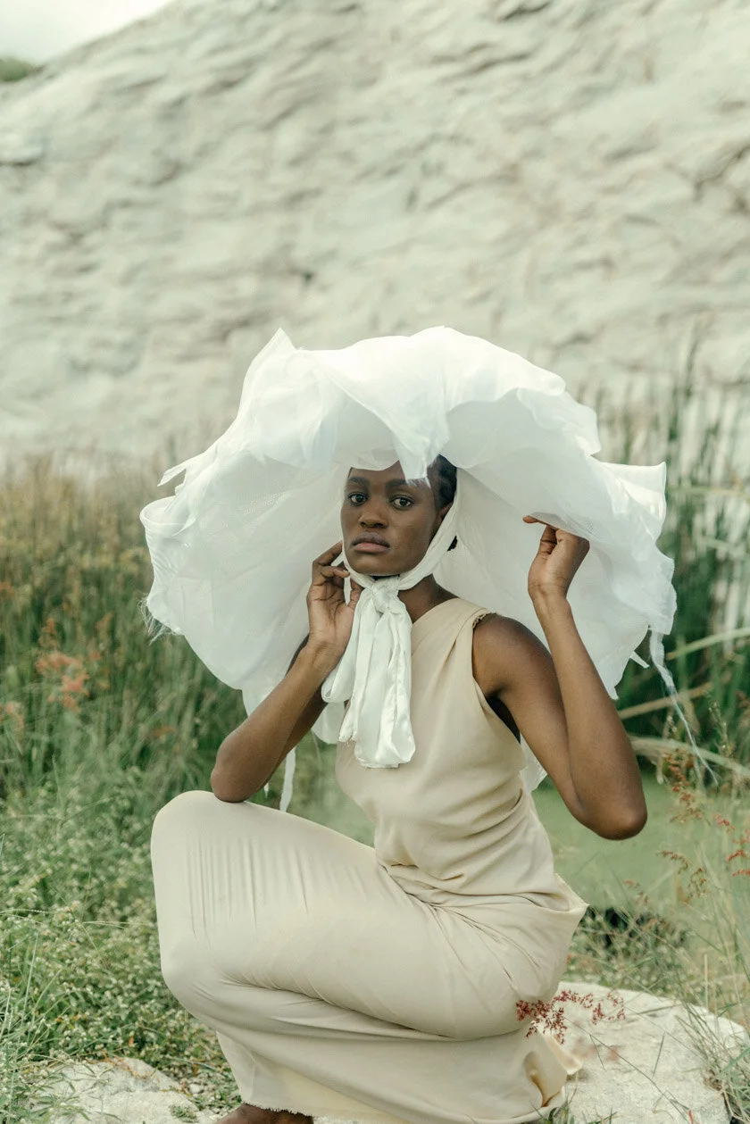 A woman in a beige dress sitting outdoors on a sandy area with green plants and a rocky background, wearing a large white hat and holding its brim while looking at the camera.