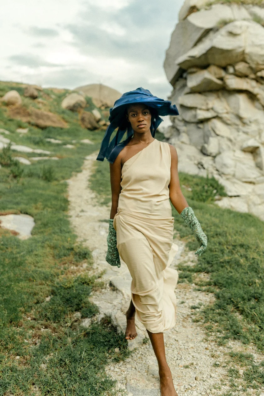 A woman walking barefoot on a rocky dirt path in a grassy, mountainous landscape, wearing a beige dress, green-patterned gloves, and a large blue hat.