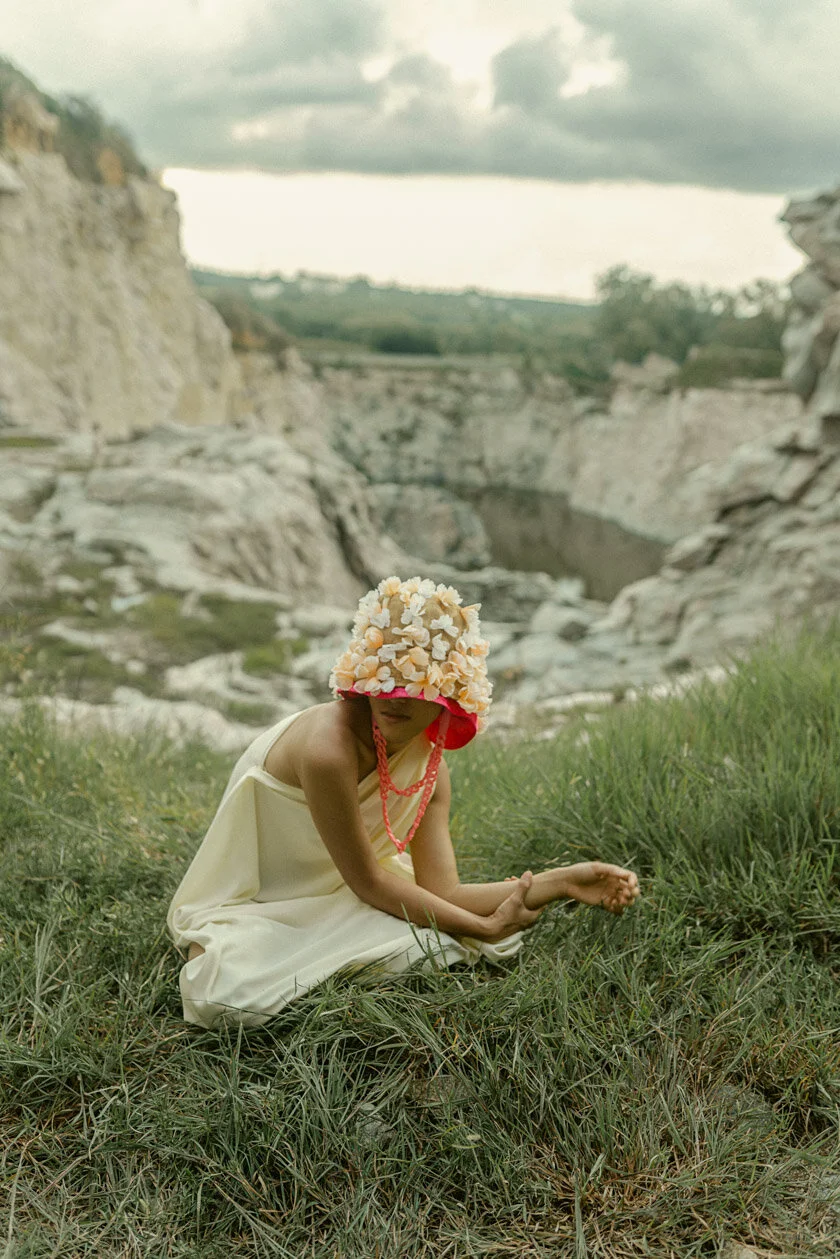 Person sitting on grass outdoors near rocky canyon, wearing a floral hat, a light-colored dress, and a red beaded necklace, with cloudy sky overhead.