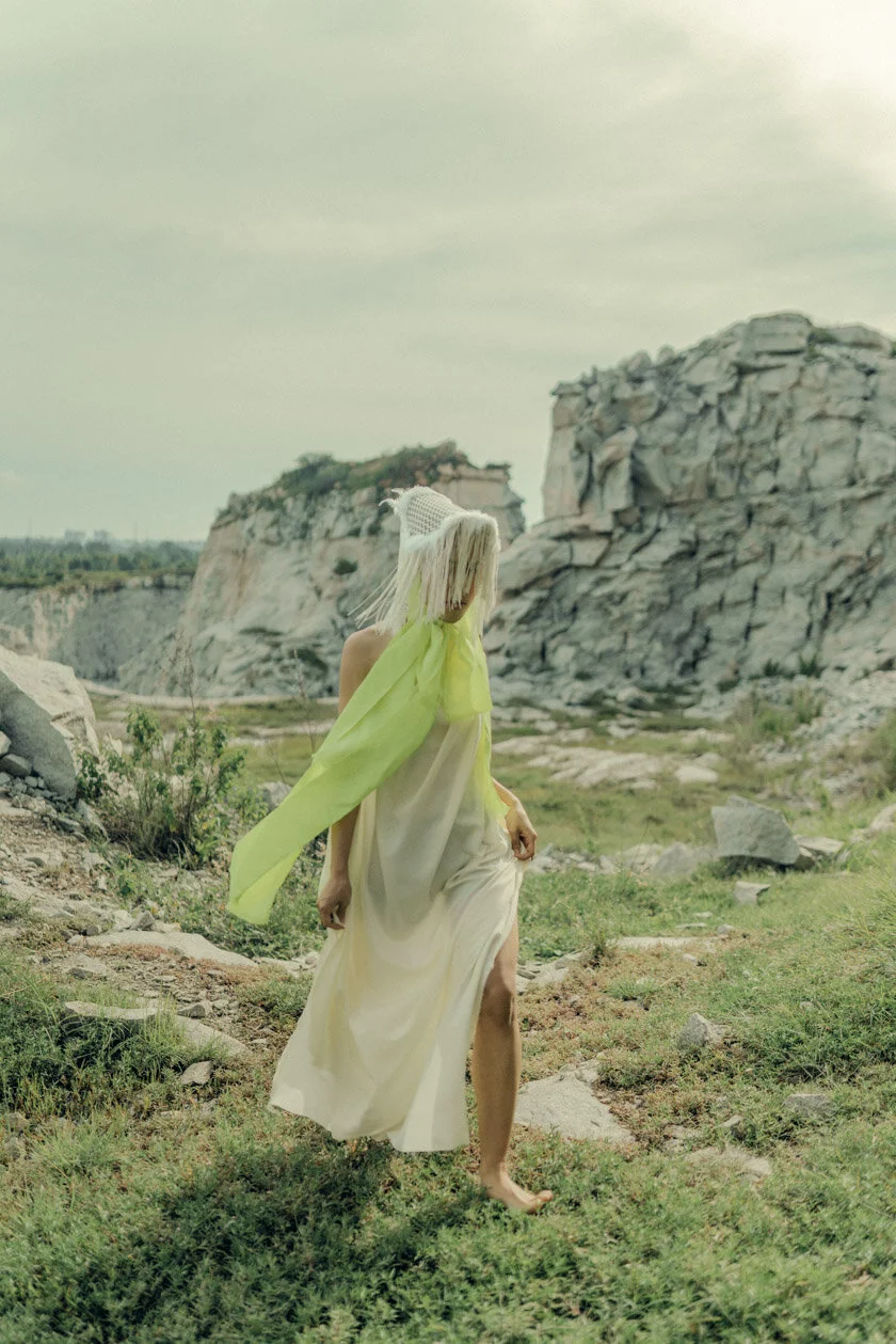 A woman with platinum blonde hair, wearing a white dress with a neon green scarf or ribbon, walking barefoot on grassy terrain with rocky cliffs in the background.