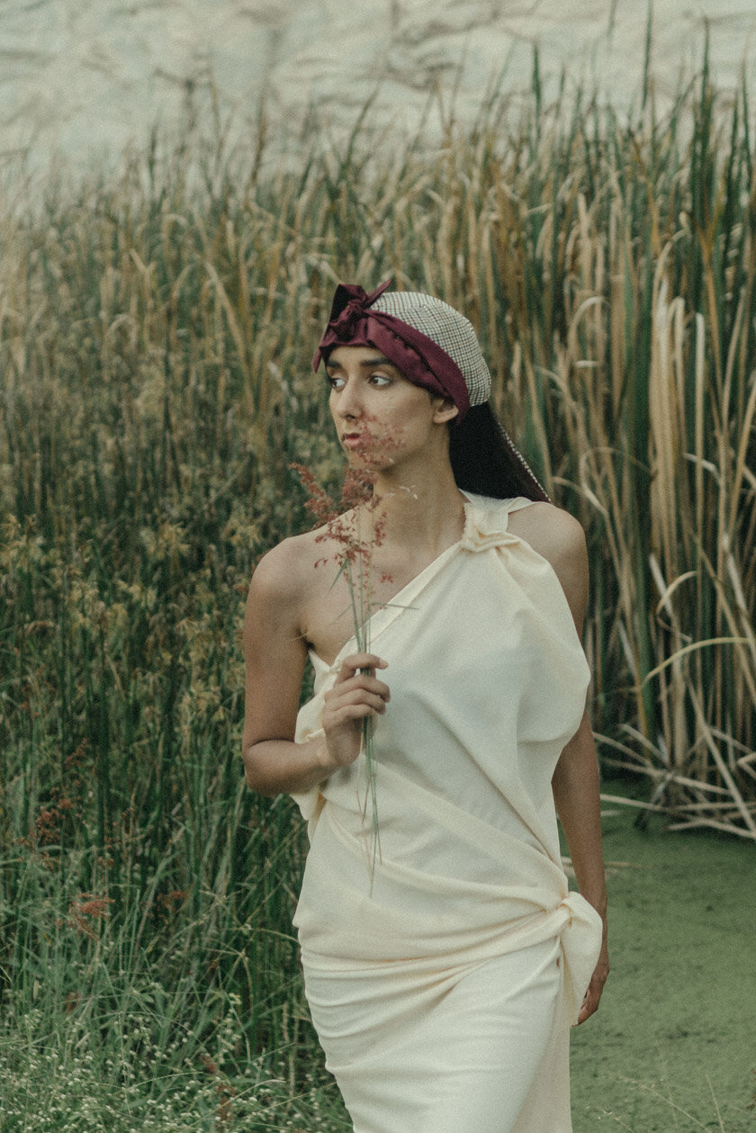 A woman dressed in a cream-colored, draped gown with one shoulder strap, standing in a field of tall grass and reeds, holding a small sprig of wildflowers near her chest, and wearing a fabric head wrap with a bow on top.