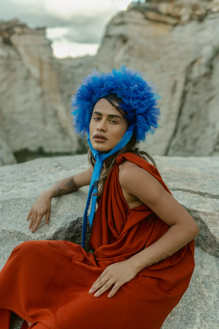 A woman in an orange dress seated on a rock, wearing a large blue fluffy hat, with a rocky mountain landscape in the background.