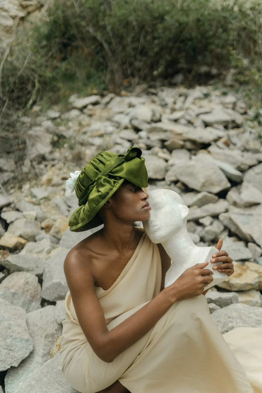 A woman wearing a beige dress and a green hat holding a white sculpture of a face in an outdoor rocky setting.