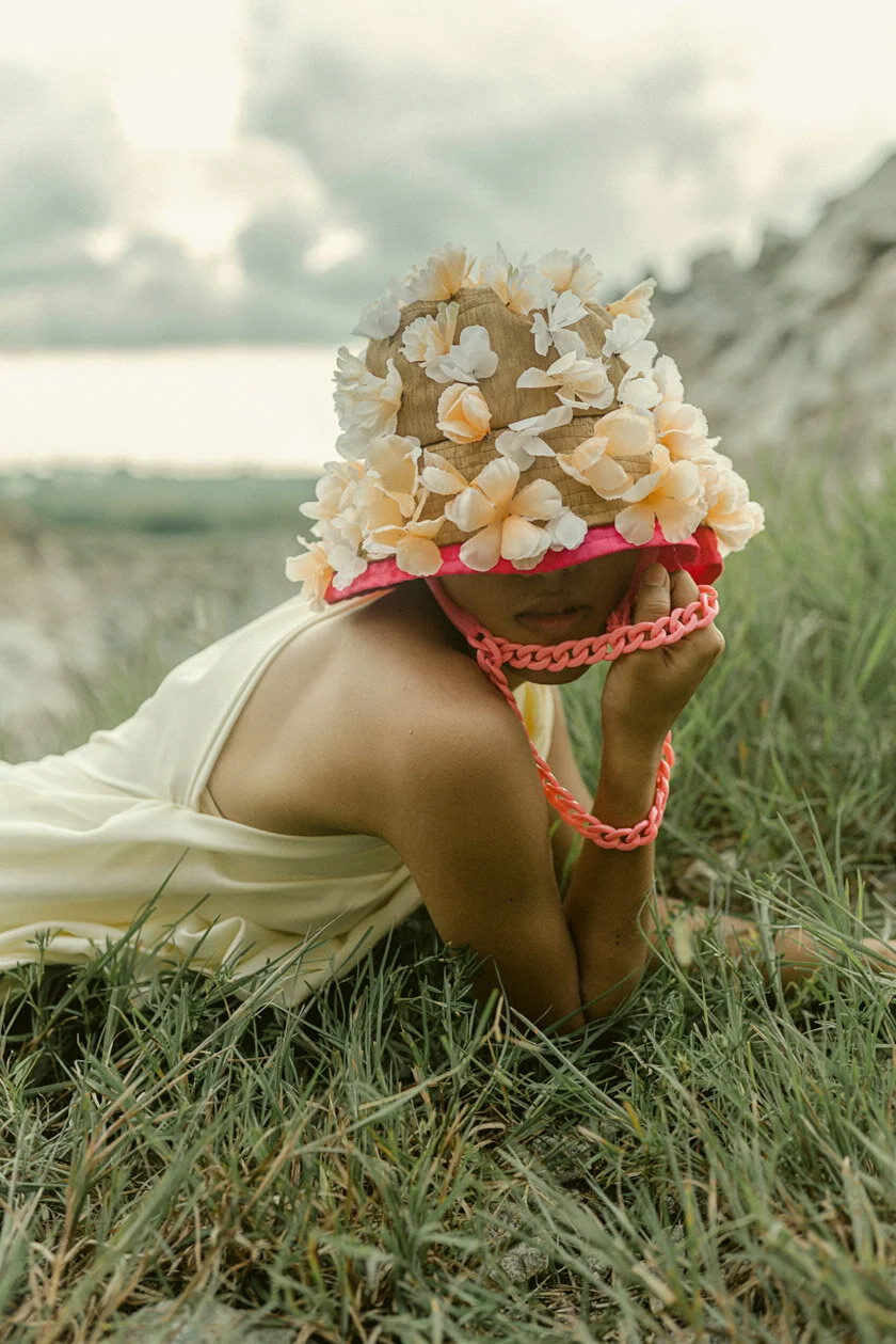 Person in a beige dress lying on grass wearing a large hat decorated with flowers and a pink chain. Gray cloudy sky in background.
