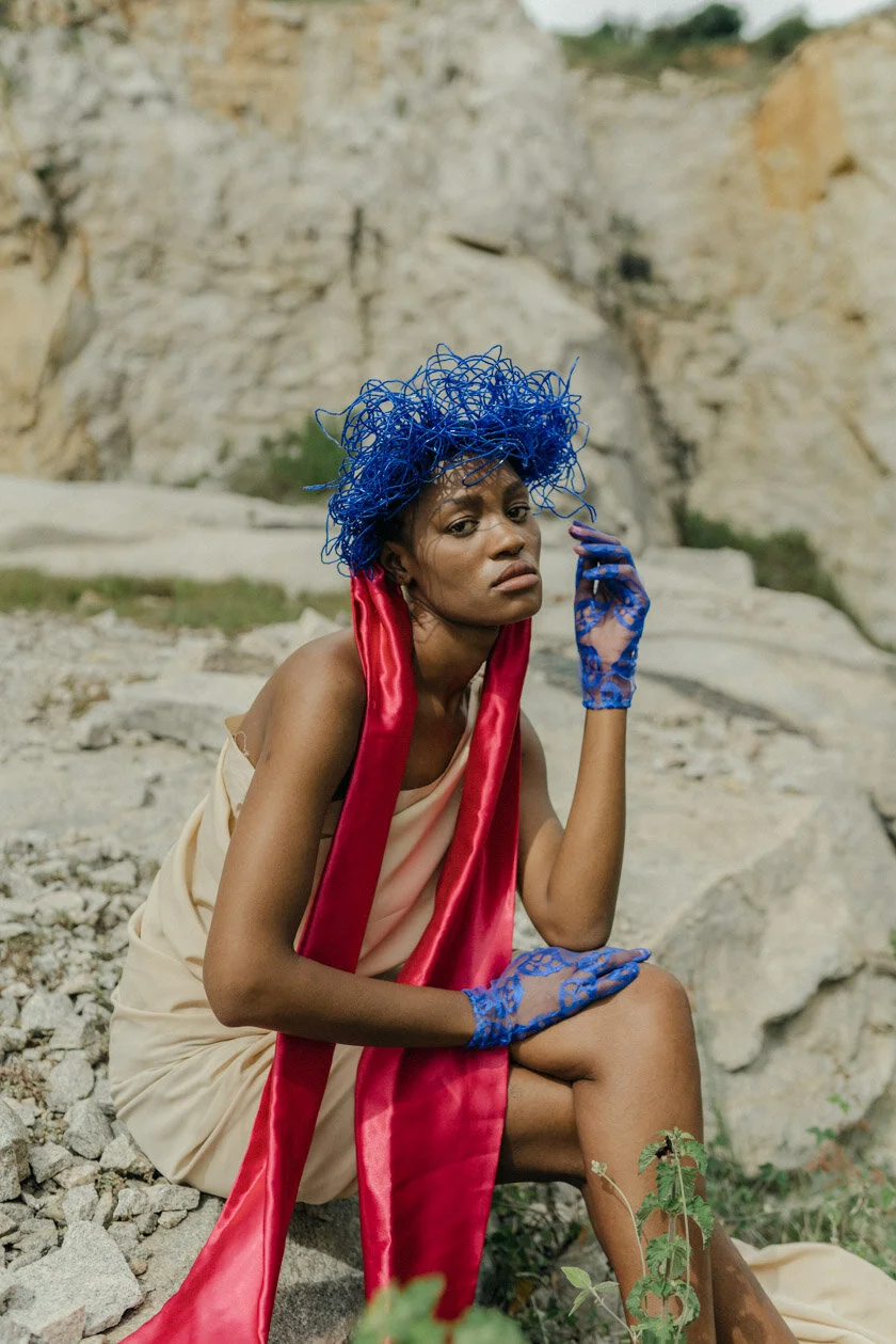 A woman with a curly blue hat and blue lace gloves sitting outdoors on rocky terrain, wearing a beige dress with a red satin scarf draped over her shoulders, near a rocky landscape.