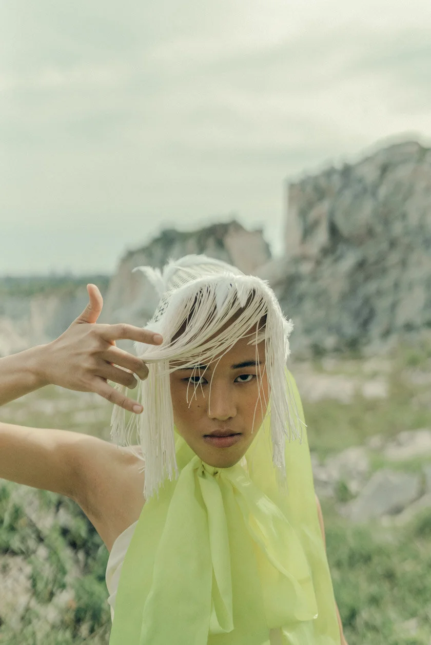 A woman with wavy platinum blonde hair posing outdoors in front of rocky cliffs, wearing a sheer yellow sleeveless top and making a gesture with her hand near her head.