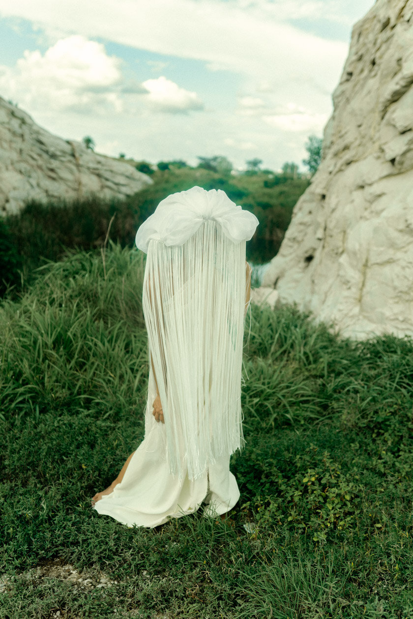 Person with long white hair and a white dress kneeling outdoors between large rocks, with a white cloth on top of their head, in a natural green landscape with grass and shrubs.
