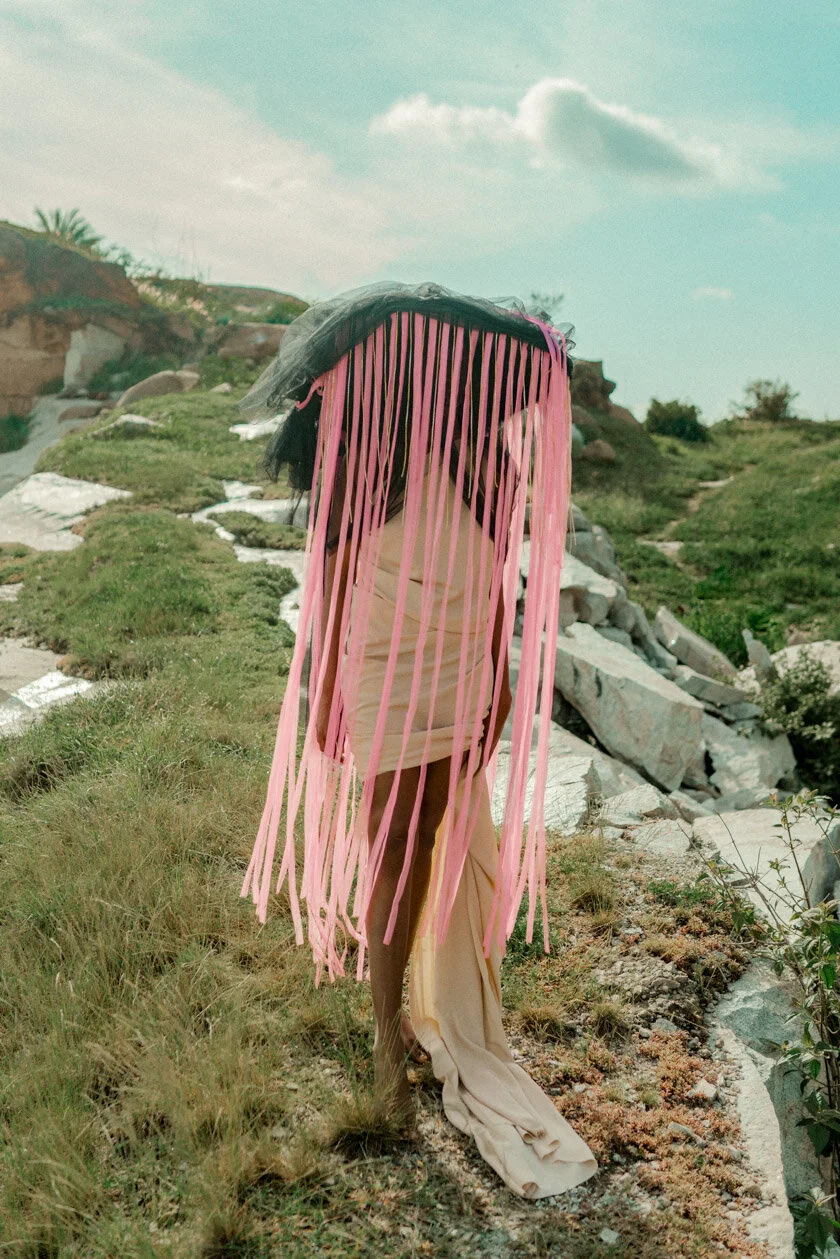 Person standing outdoors wearing a dress and a wig with long pink ribbons hanging down, with a grassy hillside and rocks in the background.
