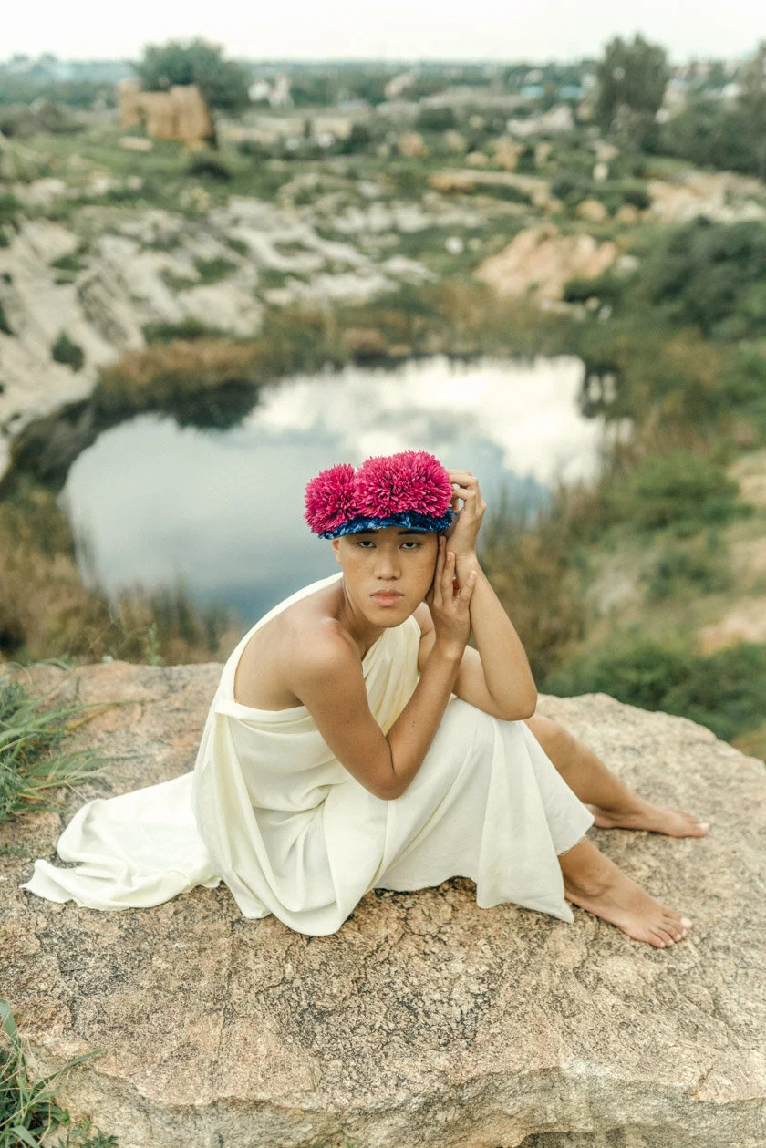 A person sitting barefoot on a rock near a small pond or lake, wearing a cream-colored dress and a crown of pink flowers, with a natural landscape and buildings in the background.