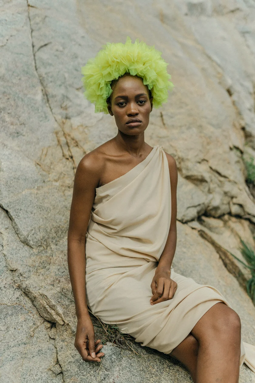 A woman sitting on rocks wearing a beige, one-shoulder draped dress and a large, bright yellow, fluffy headpiece.