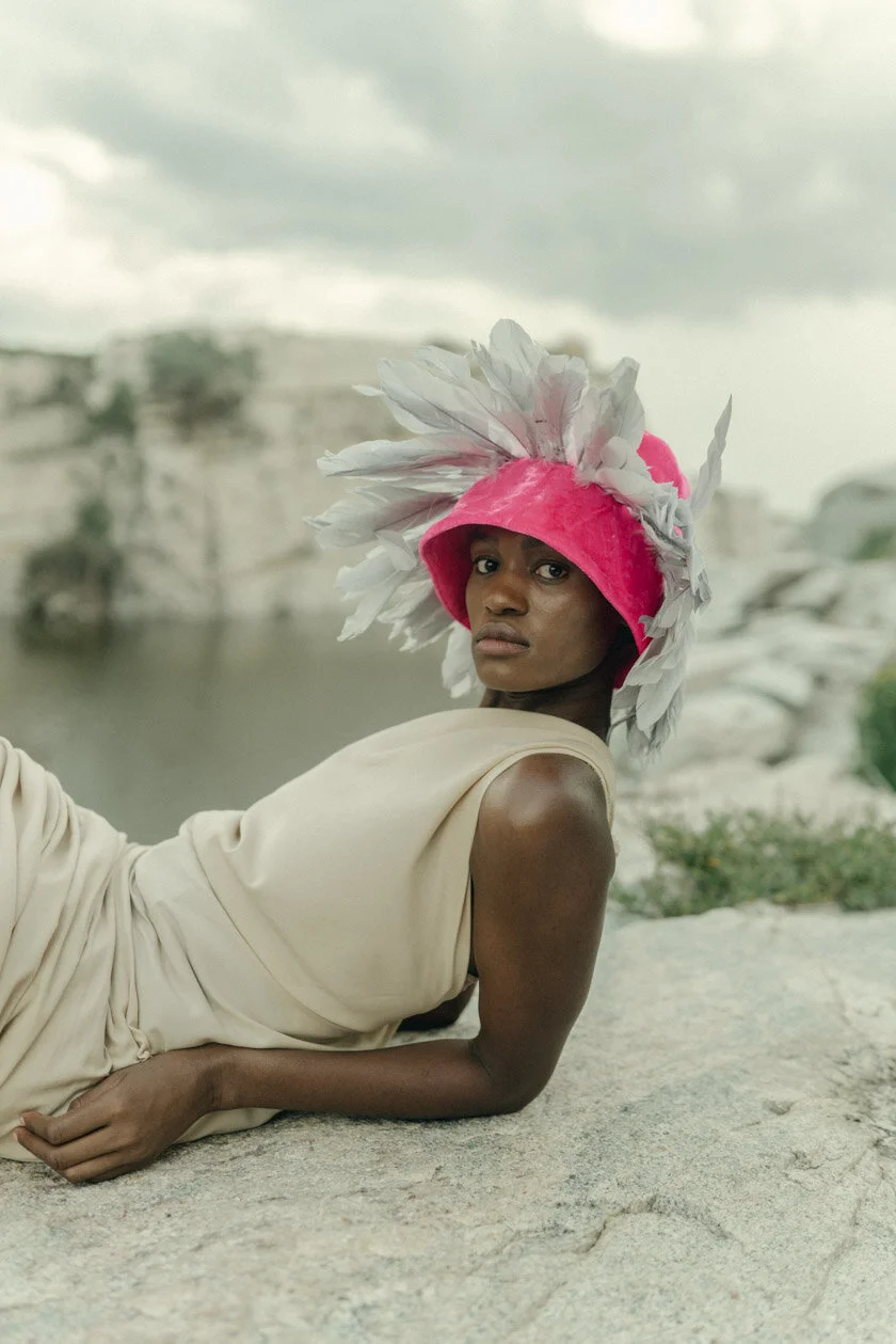 A woman wearing a bright pink hat with large white feathers, lying on a rocky surface outdoors near water with a cloudy sky in the background.
