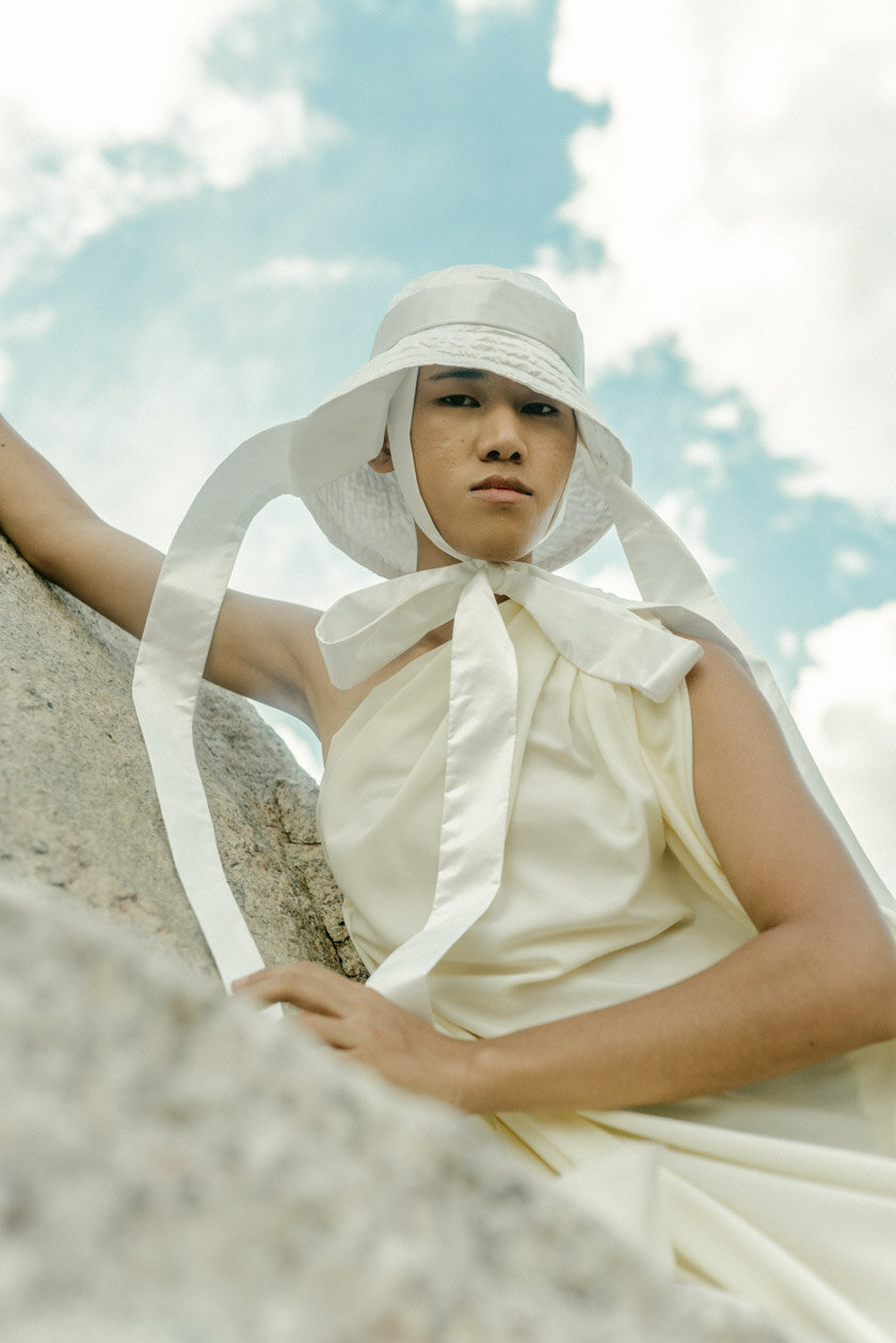 A person wearing a white sun hat and matching sleeveless dress, posing outdoors against a blue sky with clouds, leaning on a rock surface.