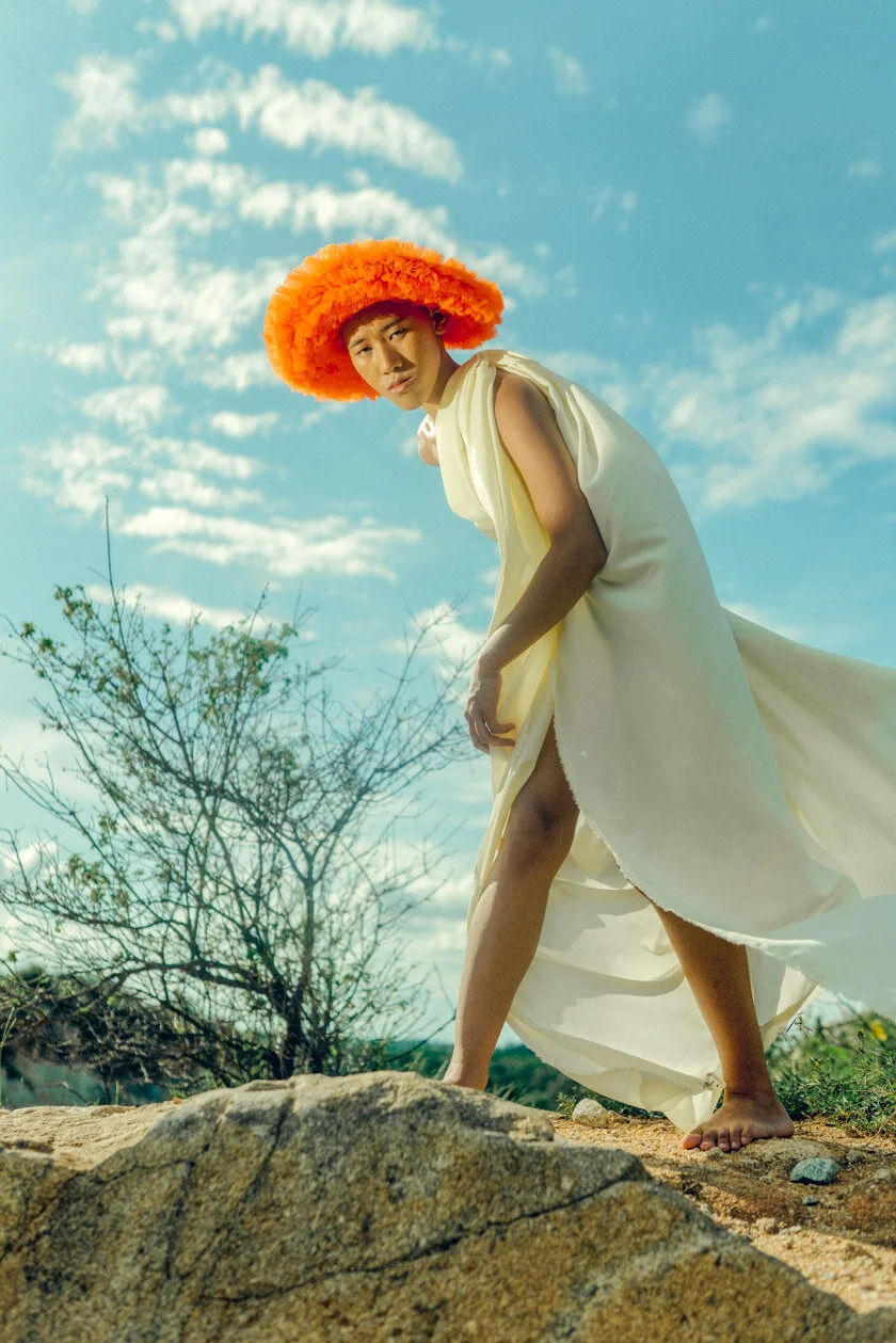 A person with fair skin and short black hair wearing a bright orange fluffy hat and a flowing pale yellow dress, standing outdoors on a rocky terrain under a partly cloudy sky.