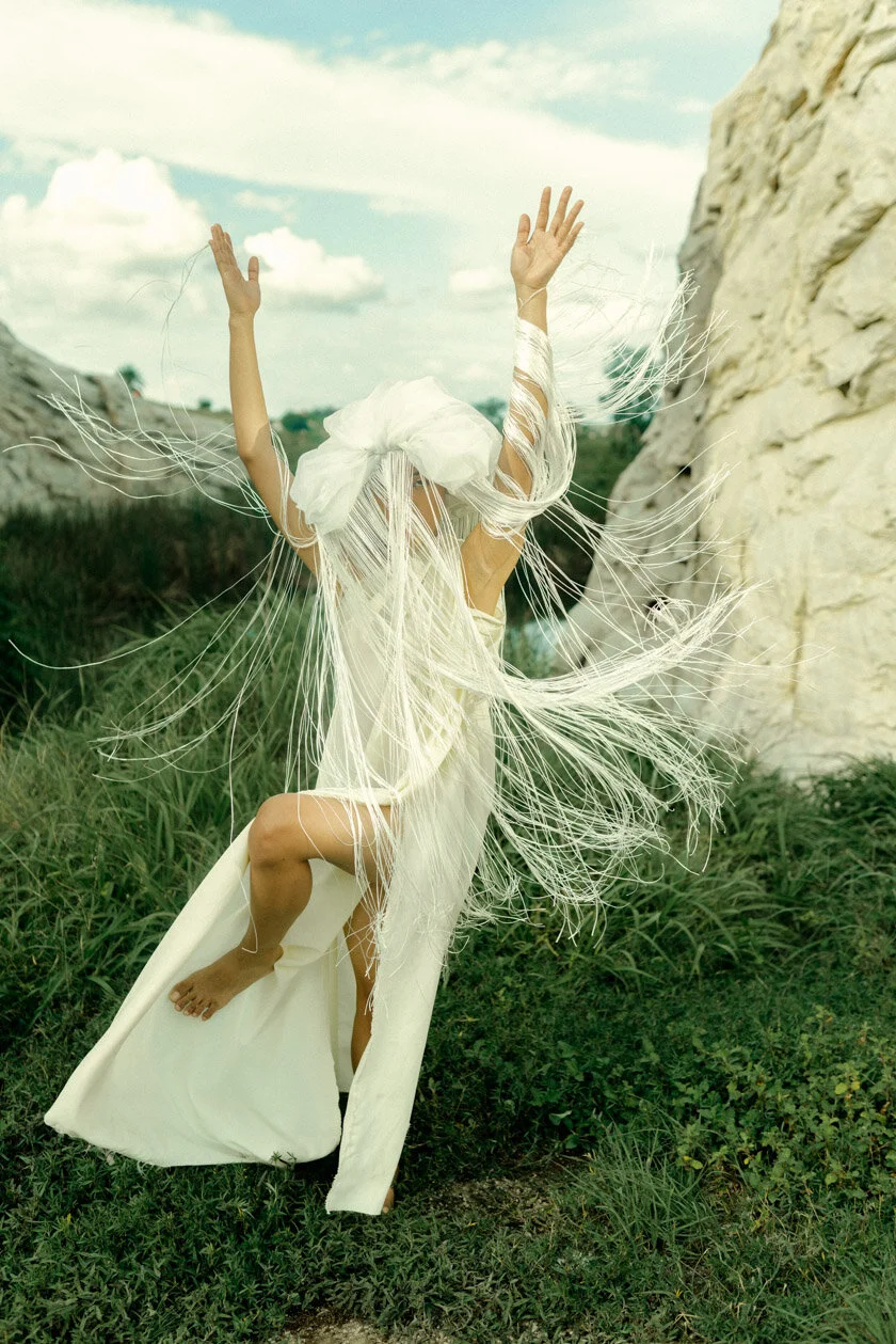 Person in a flowing white dress and head covering dancing outdoors with arms raised, standing on one foot among green grass near white rock formations under a partly cloudy sky.