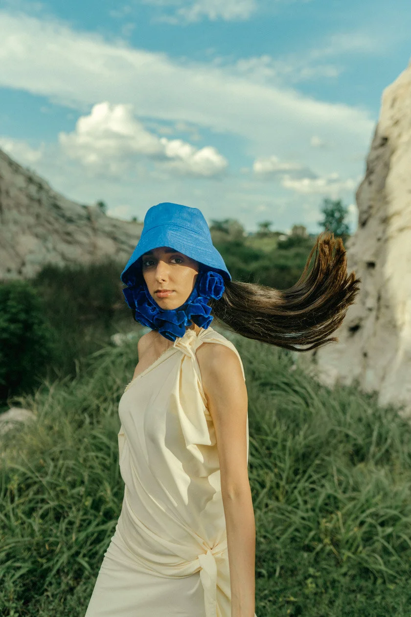 A woman with long dark hair wearing a blue bucket hat and a cream-colored dress in a grassy outdoor setting with rocky hills and a partly cloudy sky.