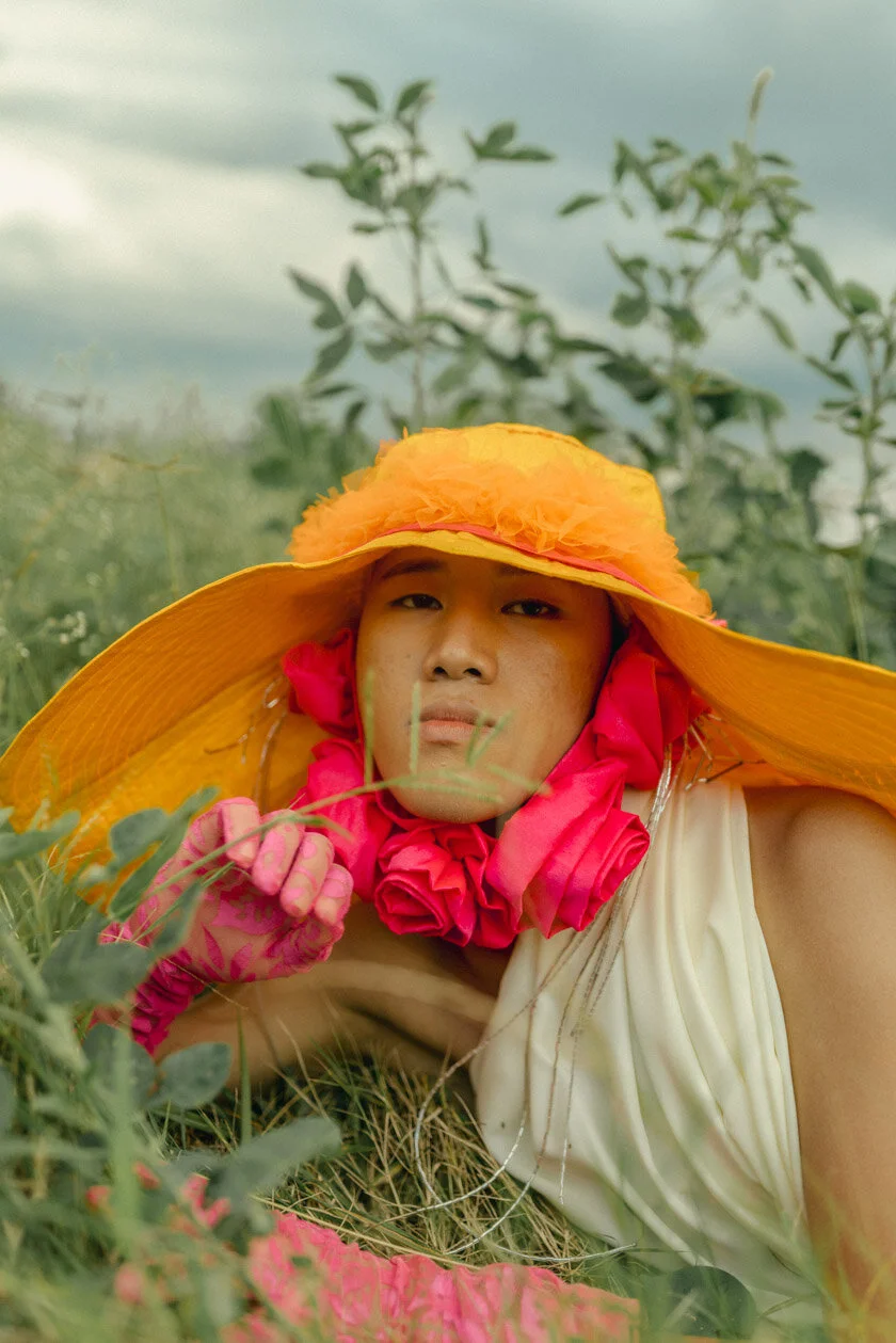 Person lying on the grass in a field, wearing a large, bright yellow hat with orange and pink accents, pink gloves, and a cream-colored dress or top, with tall green plants and a cloudy sky in the background.