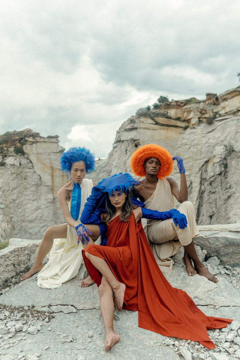 Three women dressed in colorful, dramatic costumes with bright orange and blue wigs, seated on rocky terrain in a barren, canyon-like landscape under a cloudy sky.