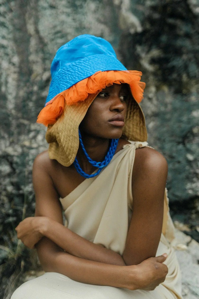 A person sitting outdoors wearing a colorful hat with orange frill, a beige draped outfit, and a blue necklace, with a rocky background.