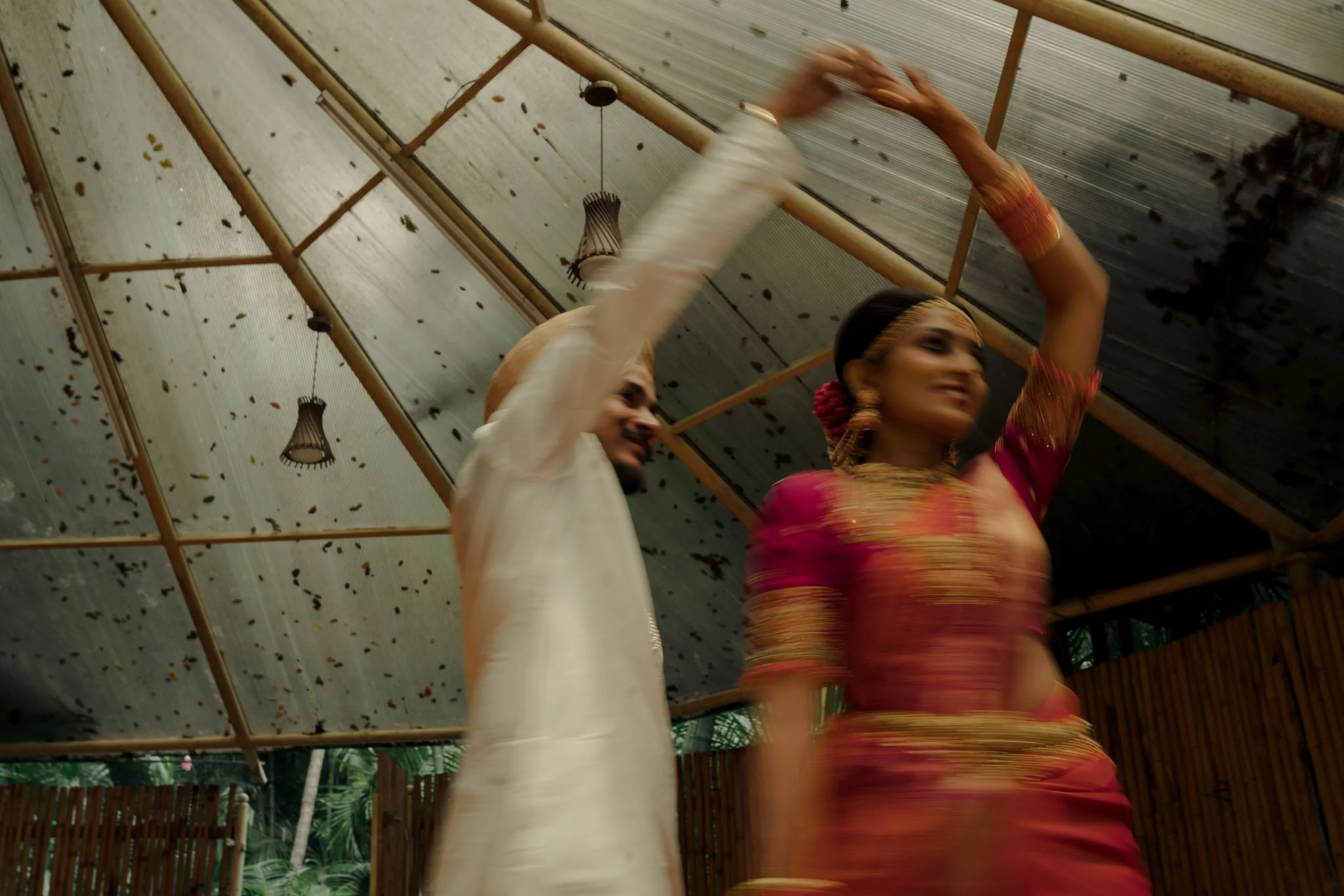 A couple dancing, with the woman in traditional Indian attire and the man in white traditional clothes, under a ceiling decorated with hanging lamps.