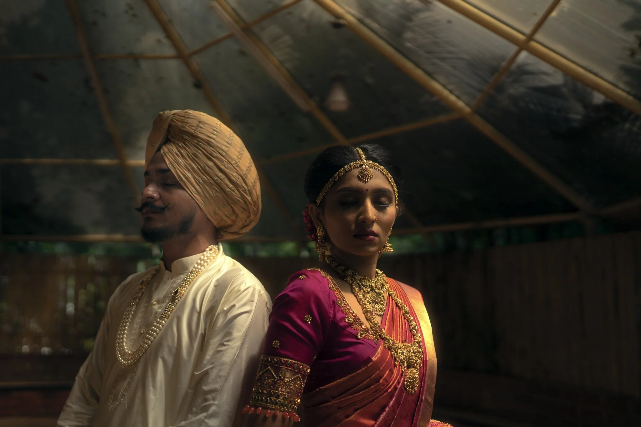 A man and woman dressed in traditional South Asian wedding attire, standing indoors under a canopy, eyes closed, appearing solemn and reflective.