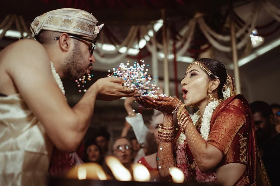 A bride in traditional Indian attire blowing colored powder from her hands towards a man in traditional attire during a wedding celebration.