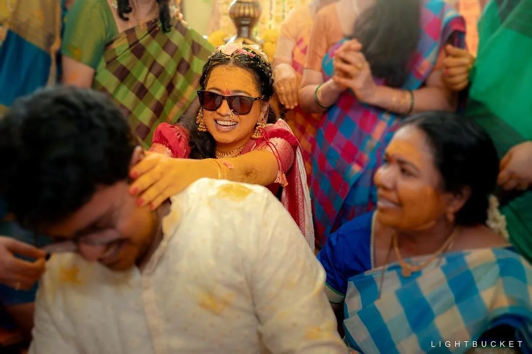 A woman with sunglasses, earrings, and a pink saree happily applying turmeric on a man's face during a celebration, surrounded by women in colorful sarees.
