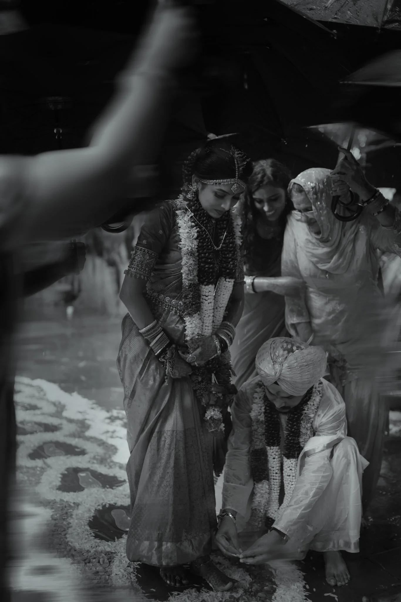 Group of women dressed in traditional Indian clothing participating in a religious or cultural festival, pouring flower petals or rice on a person lying on the ground, under an umbrella, in a street setting.