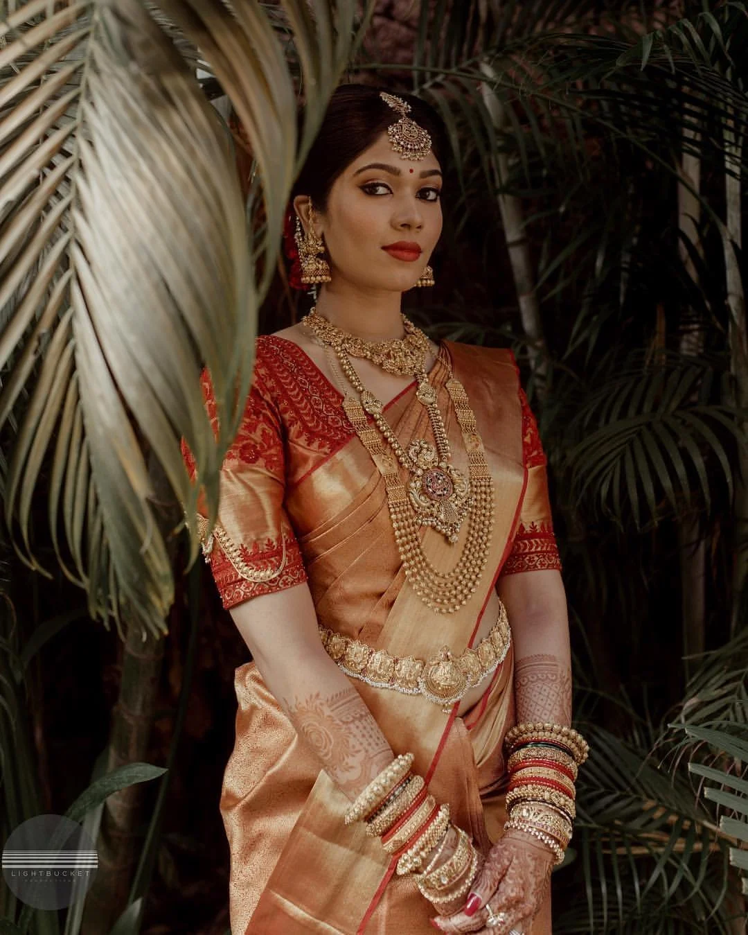A woman dressed in traditional Indian bridal attire, wearing a peach and red saree with detailed gold jewelry, including necklaces, earrings, bangles, and a maang tikka, standing amidst green foliage.