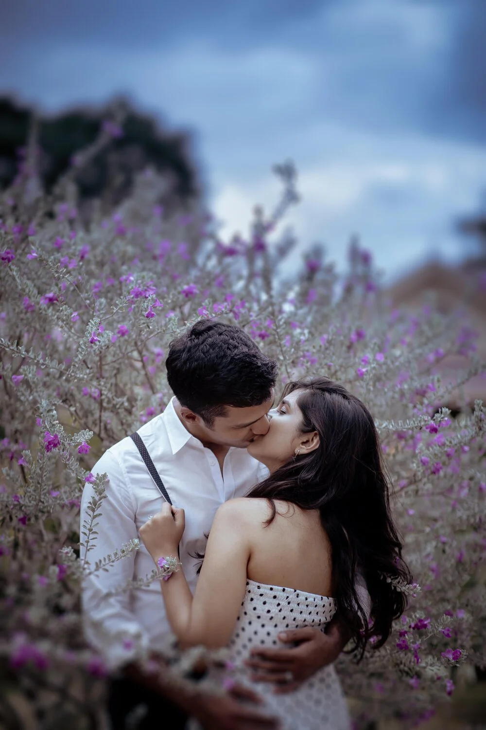 A young couple sharing a kiss in a flower field with purple blooms, in front of a cloudy sky.