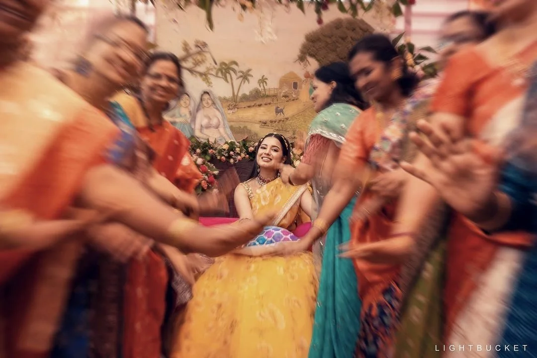 A woman in a yellow saree sitting at a celebration surrounded by women in colorful sarees, with some clapping and smiling.