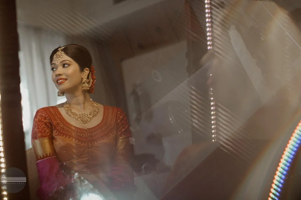 A woman dressed in traditional Indian attire, wearing jewelry, sitting indoors with a mirror reflecting her image.