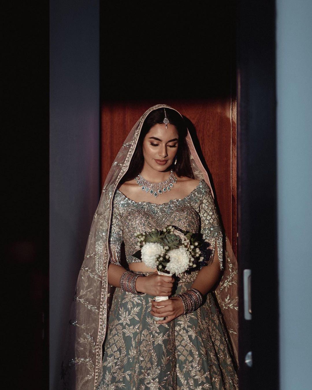 A bride in a silver and gold traditional Indian wedding dress, holding a bouquet of white flowers, standing in a doorway.