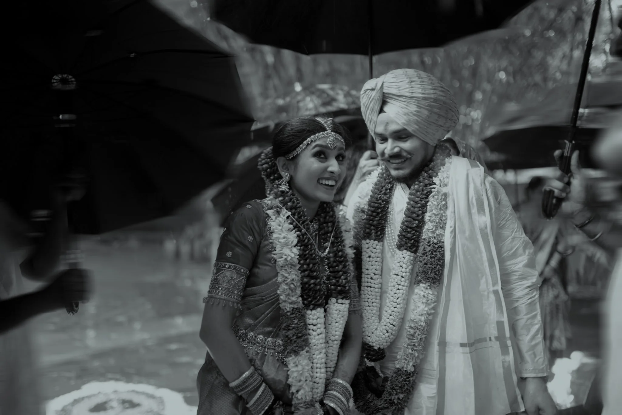 A black and white photo of a bride and groom at a wedding, smiling and looking at each other, with umbrellas over their heads.