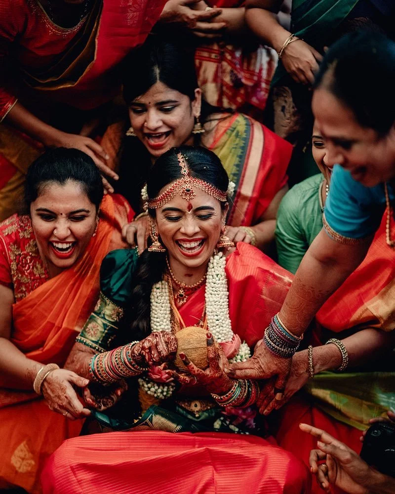 A bride in traditional Indian wedding attire, wearing jewelry and flowers, is holding a coconut surrounded by smiling women in colorful sarees during a celebration.