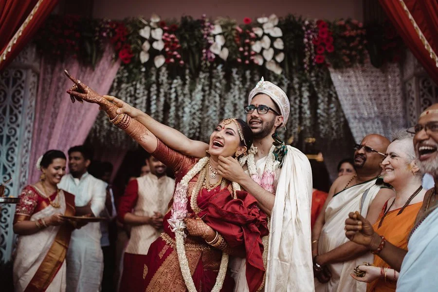 A bride and groom celebrating their wedding, with the bride pointing outward and the groom smiling, surrounded by family and guests in colorful traditional Indian attire, with a decorated floral backdrop.