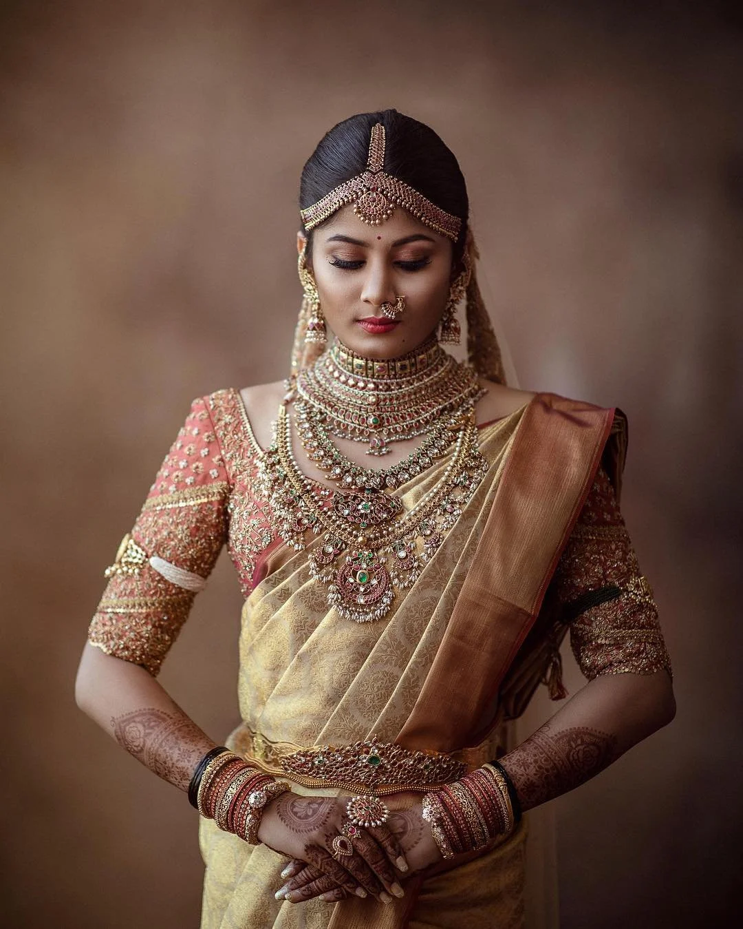 Woman in traditional Indian wedding attire with elaborate jewelry, mehndi on hands, and a sari, standing in front of a neutral background.