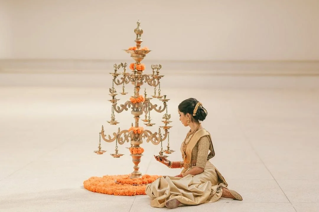 A woman dressed in traditional Indian attire sitting on the floor and lighting a brass lamp next to a large ornate ceremonial lamp with multiple tiers, decorated with orange flowers, on a plain floor.