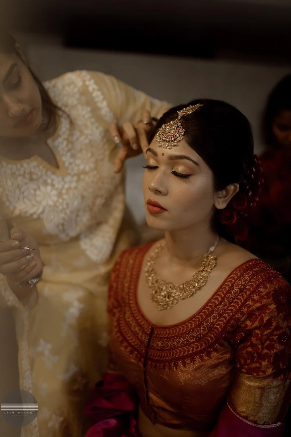 An Indian bride with traditional jewelry and makeup getting ready, possibly for a wedding, with another woman assisting her.
