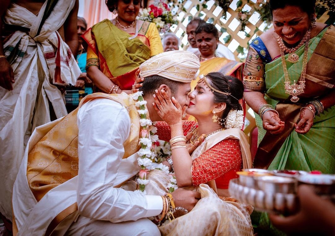 A traditional Indian wedding ceremony with the bride and groom sharing a kiss, surrounded by smiling family members dressed in colorful sarees and traditional attire.