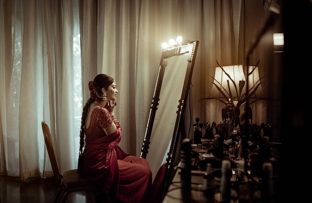 A woman dressed in traditional Indian attire, sitting in front of a mirror, in a softly lit room with cream-colored curtains and a floor lamp.