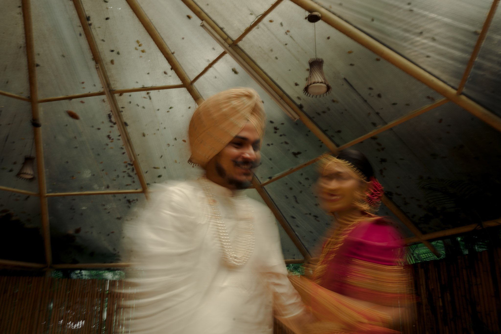 Two people smiling inside a bamboo structure with a translucent roof. The person in the foreground is wearing white traditional clothing and a turban, while the person behind is wearing a bright pink outfit.