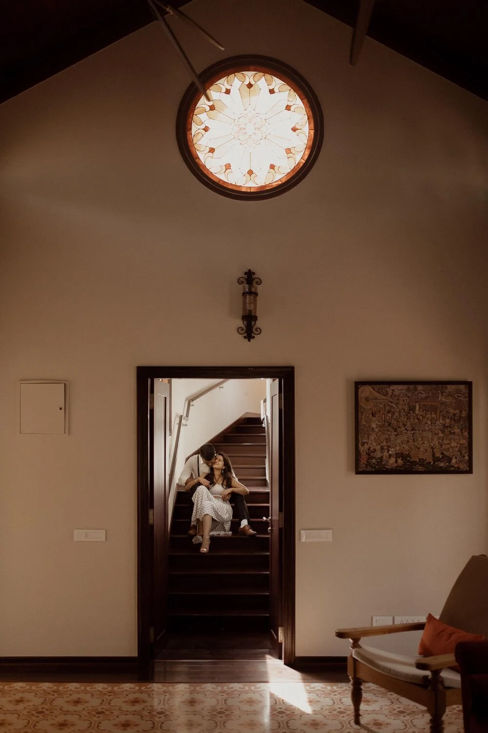 A woman sitting on stairs with a man kissing her forehead in a warmly lit interior. The staircase is viewed through an open doorway, with a decorative stained glass window above.