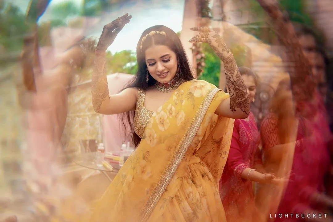 Young woman in a yellow floral saree dancing at an outdoor celebration, surrounded by women in pink and red traditional dresses.