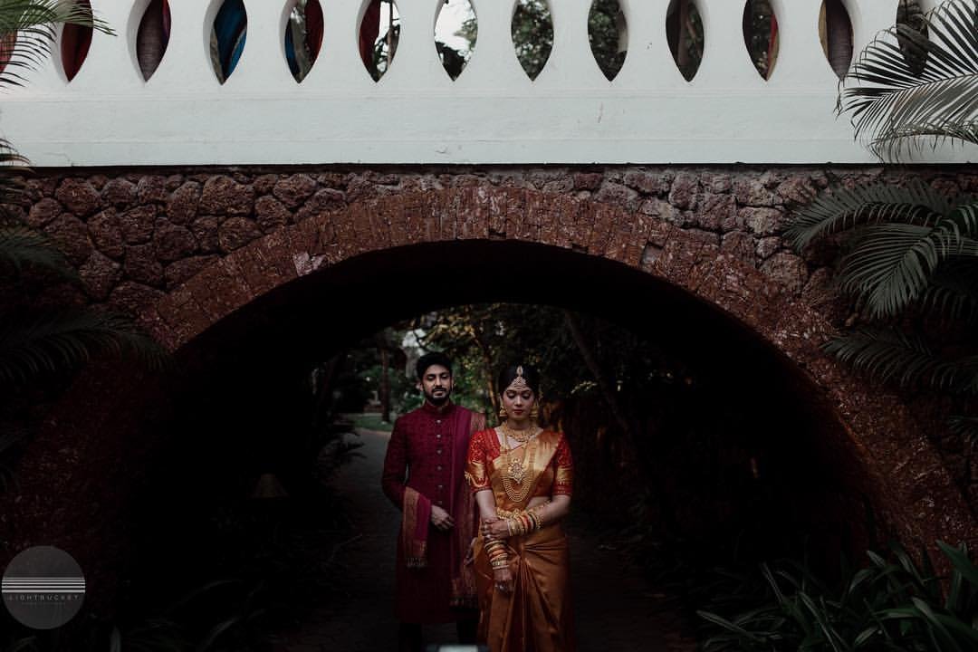 A couple dressed in traditional Indian wedding attire standing under a brick archway surrounded by lush green plants.