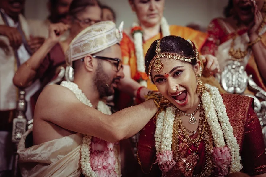 Indian bride and groom at their wedding, with the groom playfully grabbing the bride's cheek, surrounded by family members.