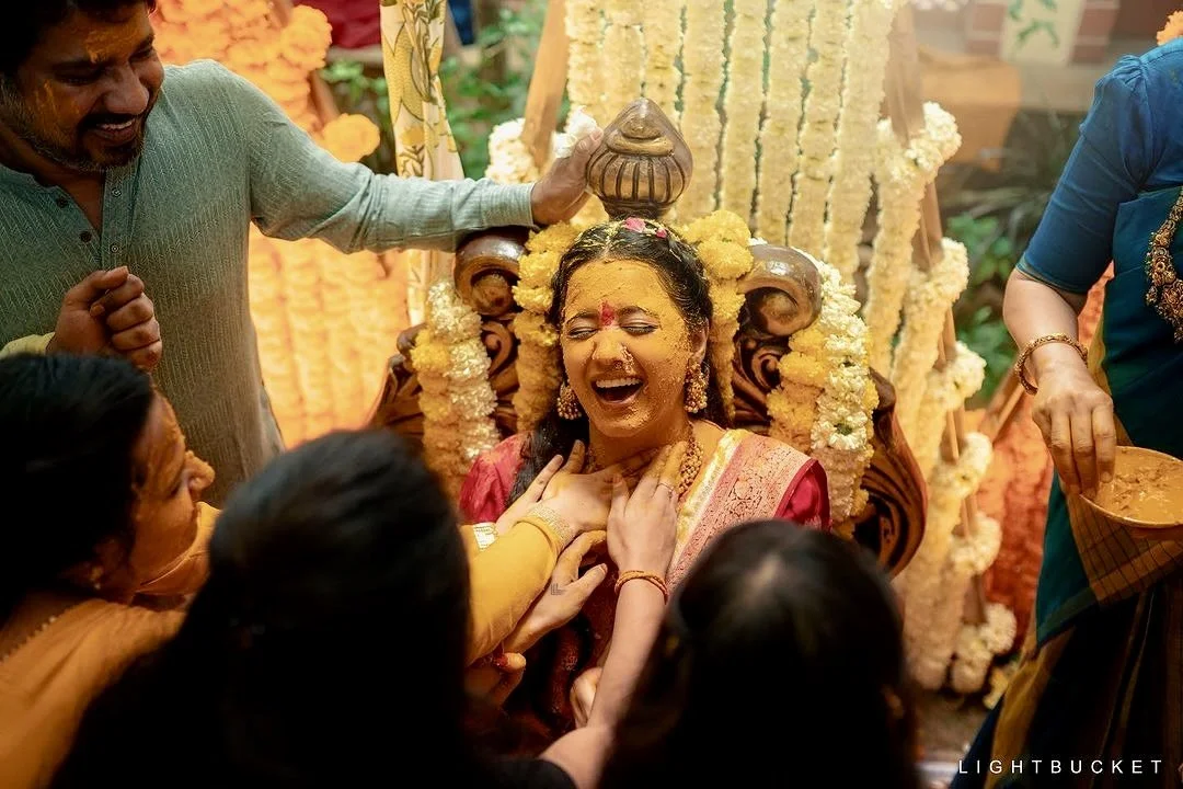 A woman dressed in traditional Indian attire, seated on an ornate chair with floral decorations, laughing joyfully during a religious or cultural ceremony surrounded by others helping her celebrate.